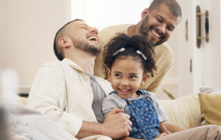 Two men and a young girl sit on a couch, smiling and laughing together in a warmly lit living room.