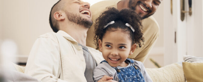 Two men and a young girl sit on a couch, smiling and laughing together in a warmly lit living room.