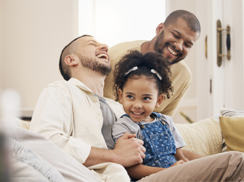 About The Adoption Projects Two men and a young girl sit on a couch, smiling and laughing together in a warmly lit living room.