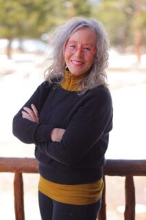 Older woman with gray hair and red glasses stands outdoors, smiling with arms crossed, wearing a black sweater over a mustard yellow top. Trees and snow are visible in the background.