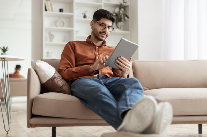Man sitting on a beige sofa, holding and using a tablet device, with legs stretched out and a relaxed posture in a bright living room.