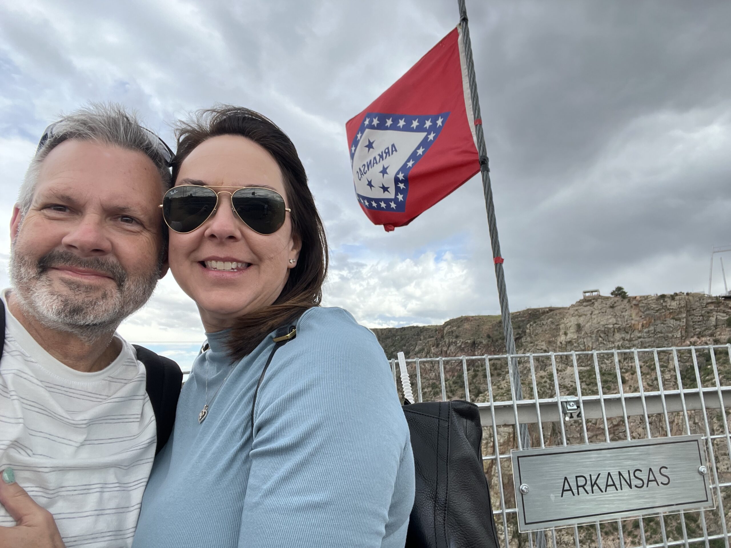 A man and woman stand in front of a fence with an "Arkansas" sign and a waving Arkansas state flag on a cloudy day.