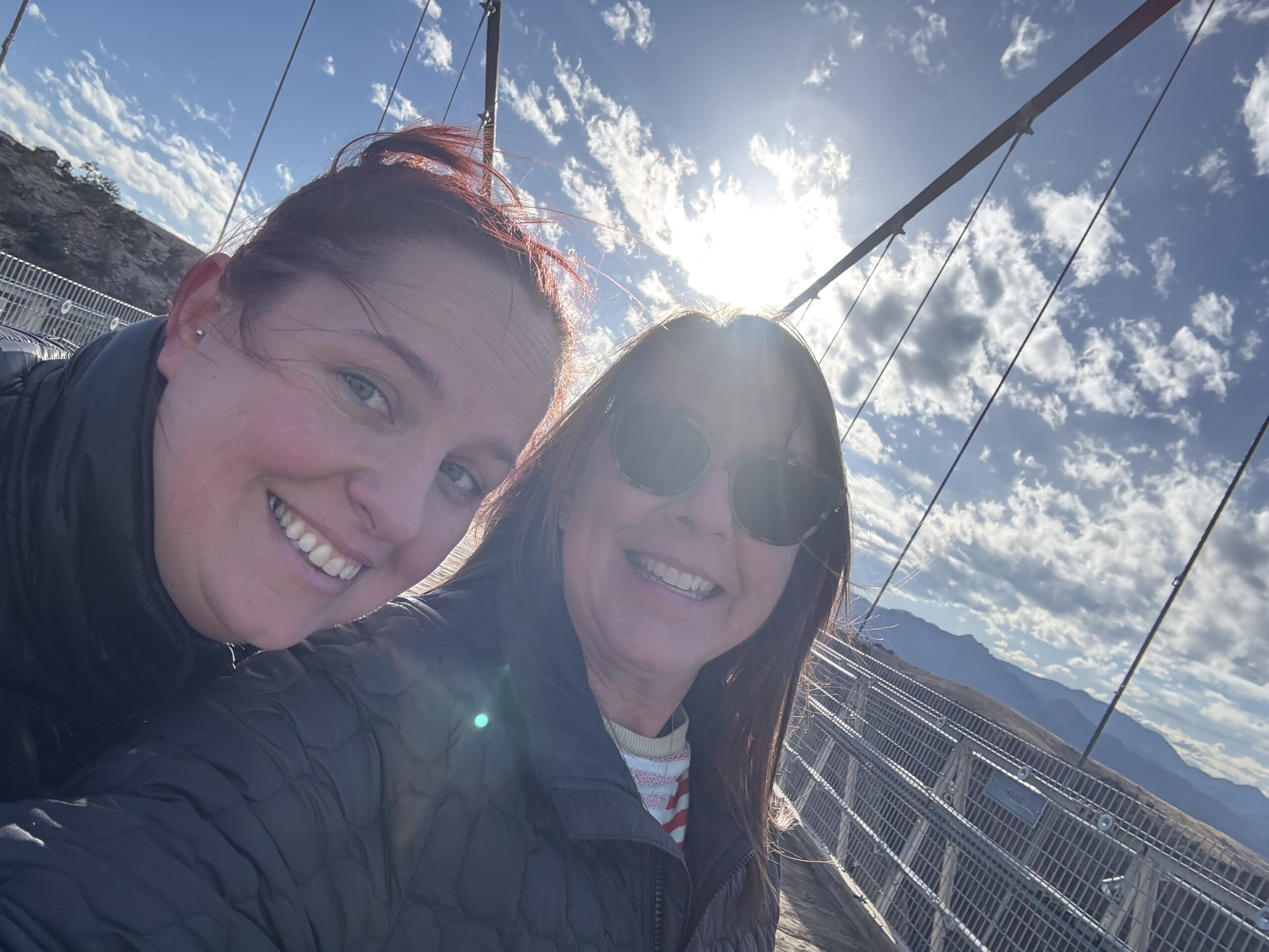Two women in jackets smile for a selfie on a suspension bridge with a bright sun and partly cloudy sky in the background.