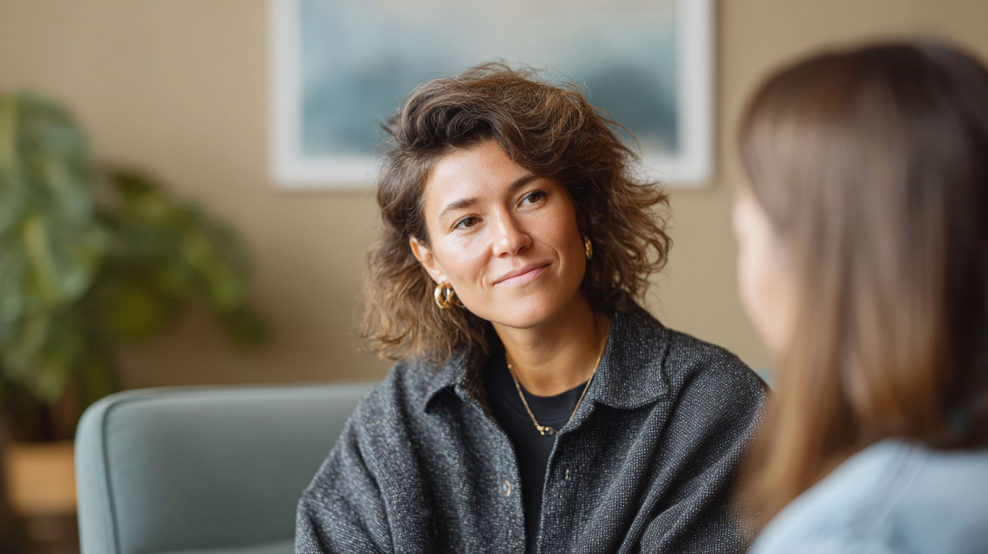Shop with Us A woman with wavy brown hair and gold hoop earrings listens attentively to another person in an indoor setting.