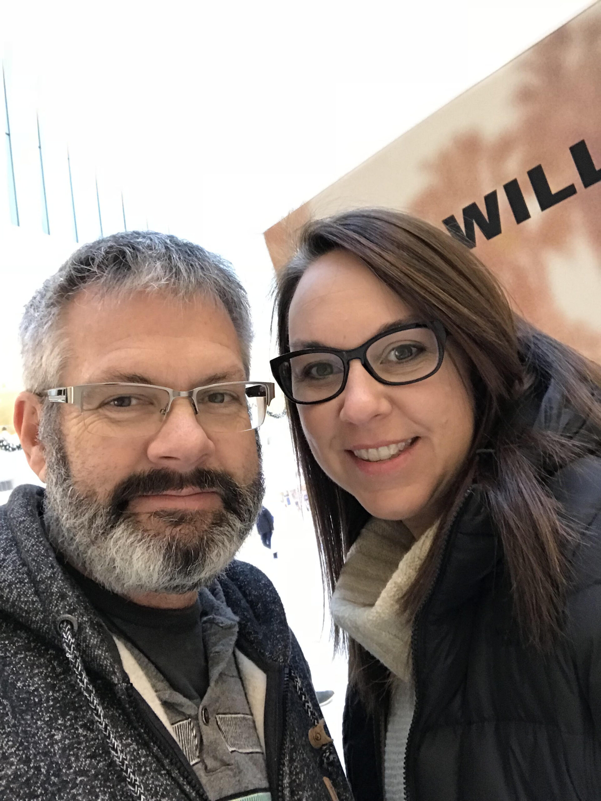 TAP – Our Story Two people wearing glasses stand close together indoors, smiling at the camera. The background includes a partially visible sign with bold black letters.